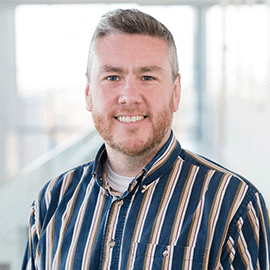 A man wearing a striped blue shirt smiles at the camera in front of a large window.